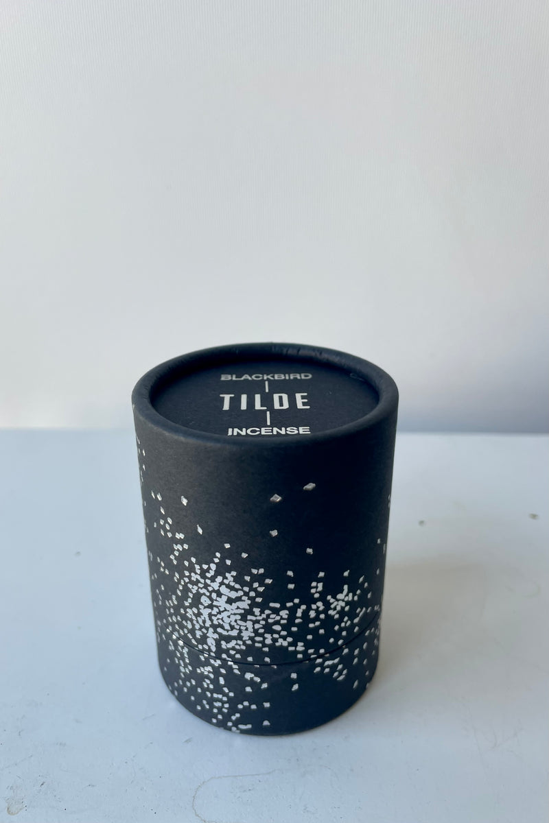 A cylinder of Blackbird incense sits on a white table in a white room. The incense is called Tilde. The packaging features a black background with silver detail of clusters of small irregular geometric shapes.