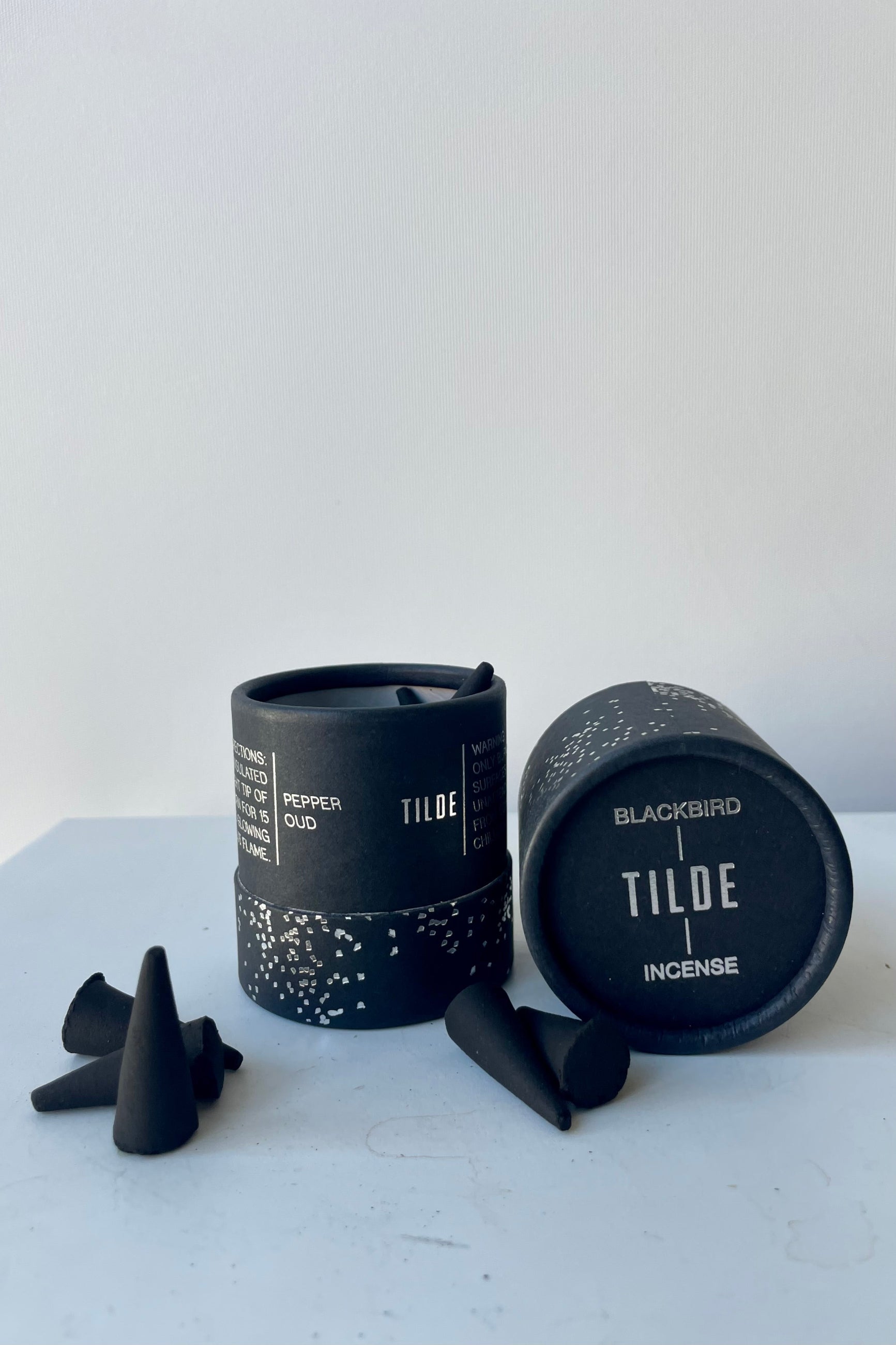 A cylinder of Blackbird incense sits on a white table in a white room. The incense is called Tilde. The packaging features a black background with silver detail of clusters of small irregular geometric shapes. To the front and right sits a scattering of black incense cones. ©Sprout home