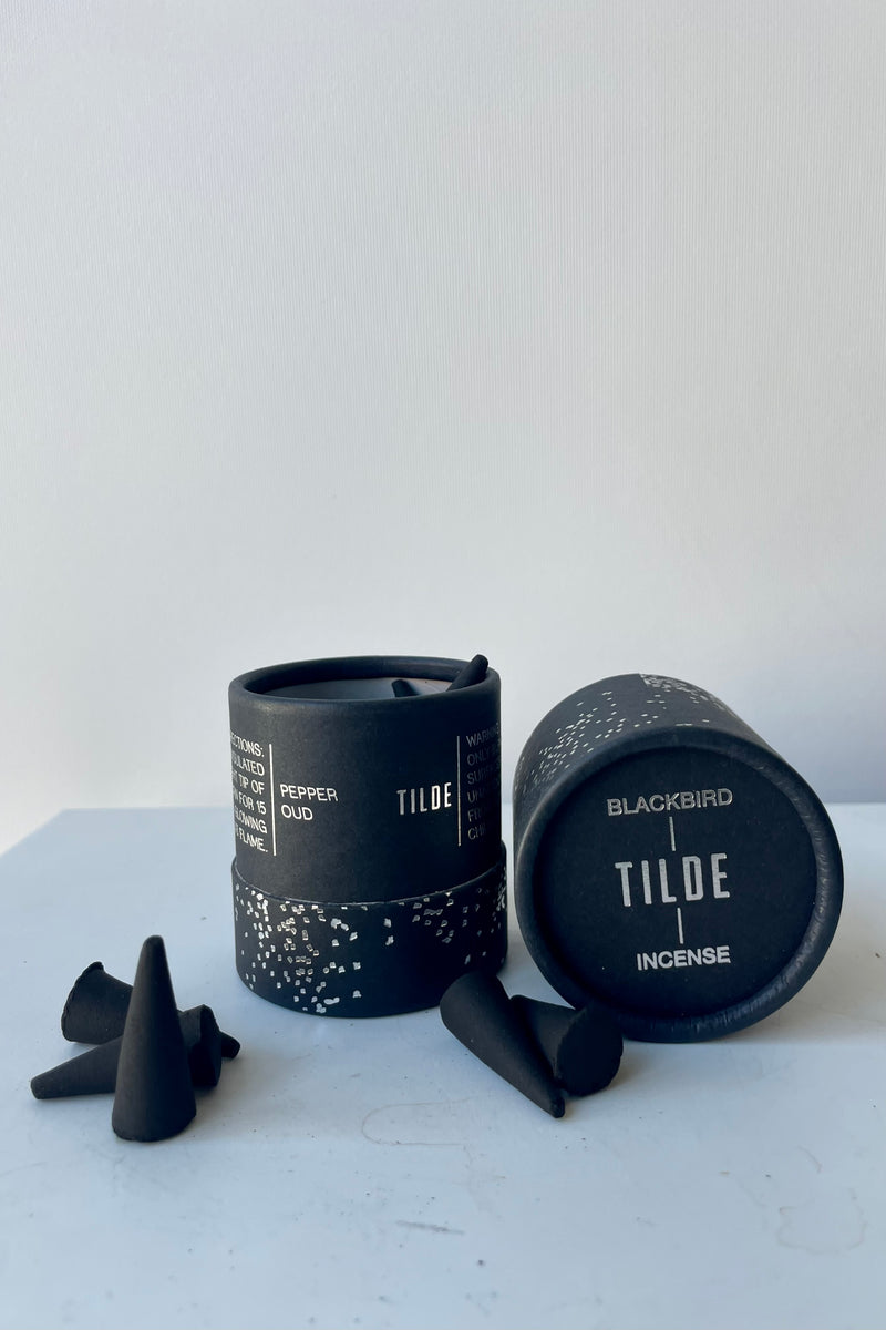 A cylinder of Blackbird incense sits on a white table in a white room. The incense is called Tilde. The packaging features a black background with silver detail of clusters of small irregular geometric shapes. To the front and right sits a scattering of black incense cones.