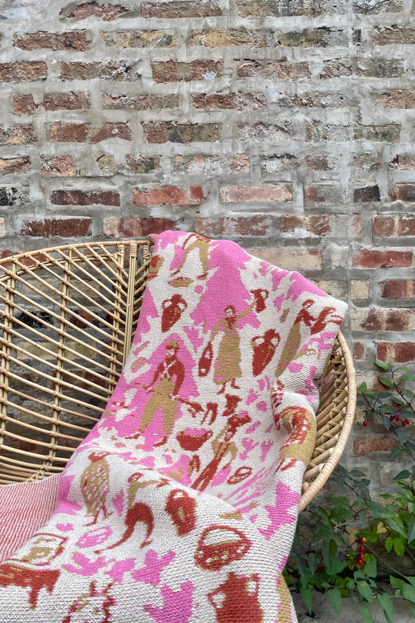 Photo of pink and tan pattern of Swiss Fields Carnation blanket on a rattan chair against a brick wall.