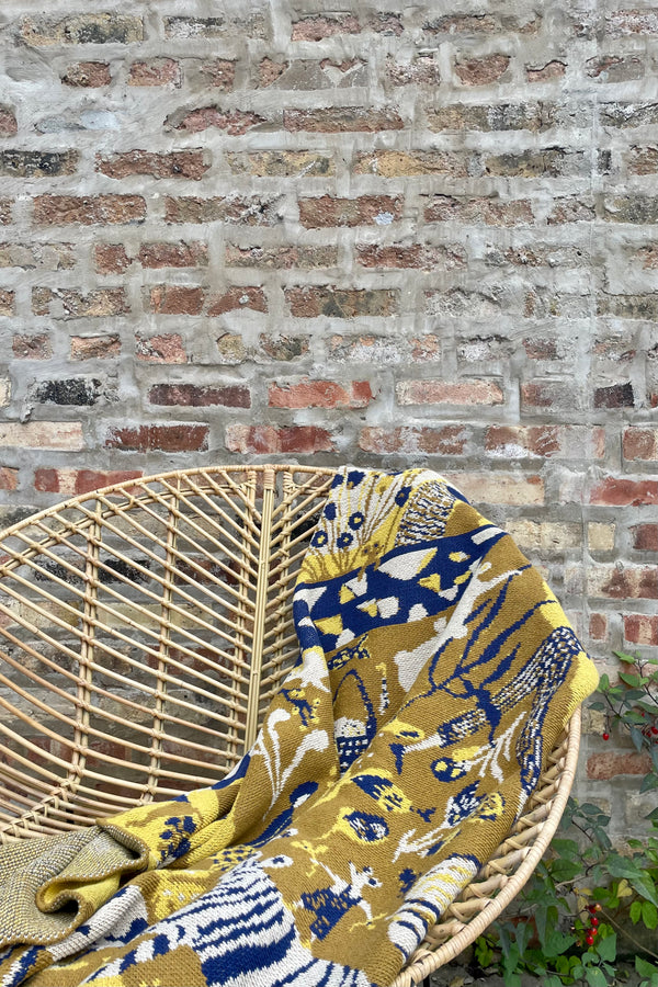 Photo of the golden and blue color pattern of Swlls Ochre blanket on a rattan chair against a brick wall