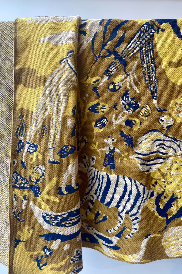 Photo of Swells Ochre woven blankiet with golden and blue patterns against a white wall.