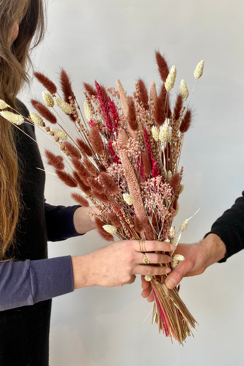 Photo of hands exchanging Blind Date preserved floral arrangement in front of a white wall.