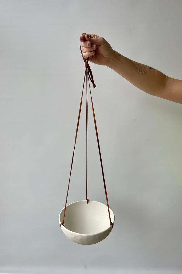 Photo of a hand holding a porcelain classic hanging planter with leather cord against a white wall looking down.
