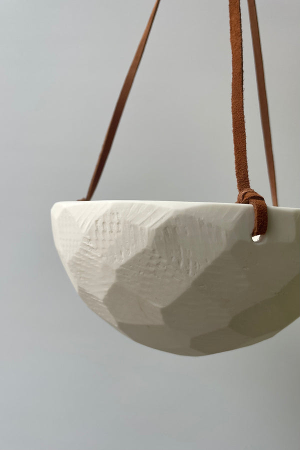 Close photo of detailed texture of a porcelain classic white hanging planter and leather cord against a white wall.