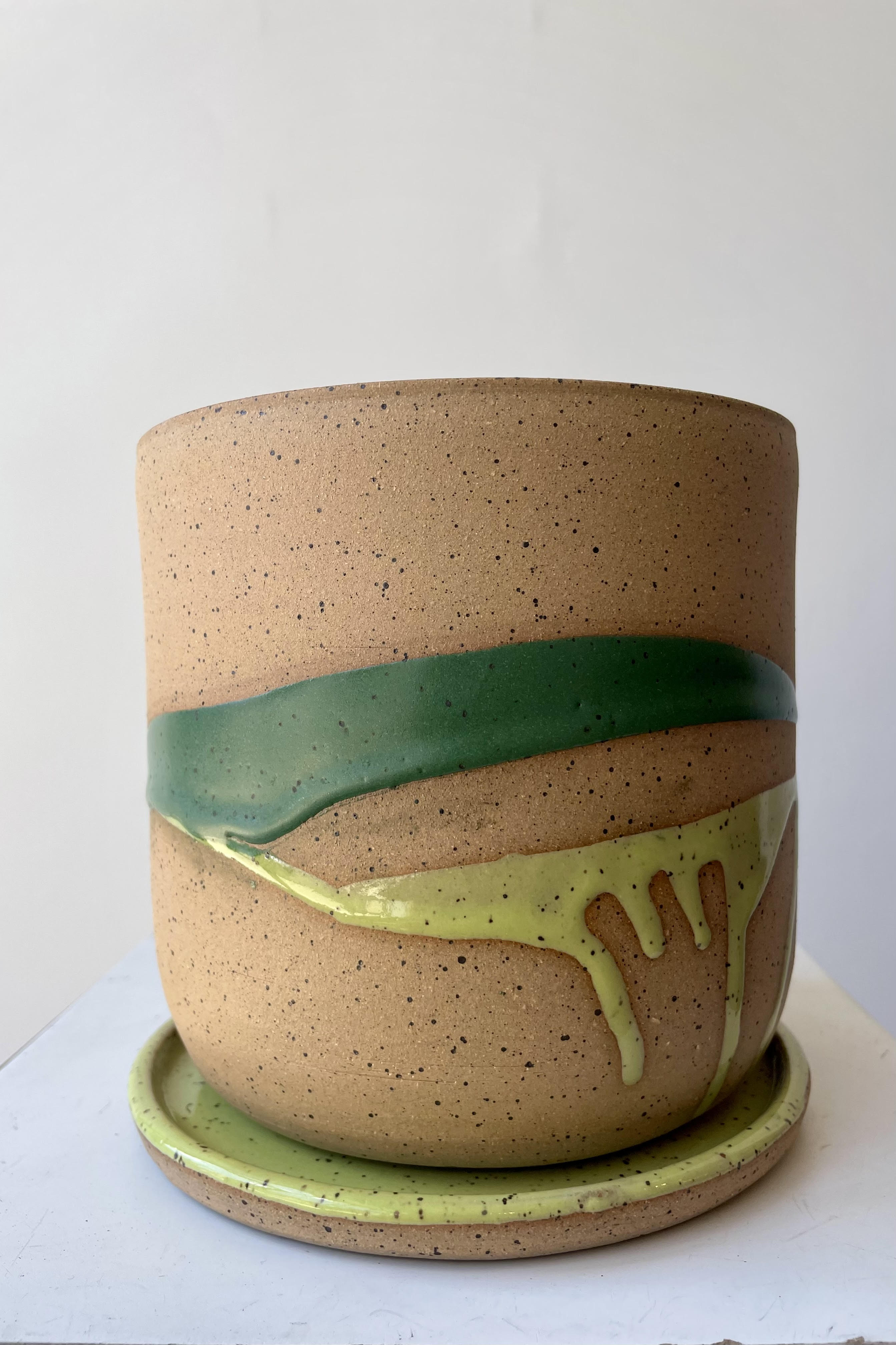 A cylinder plant pot on a saucer. The pot is made of beige clay with a darker speckle pattern. There is a semi-gloss glaze of two shades of green dripped across the planter. The pot is photographed in a white room.