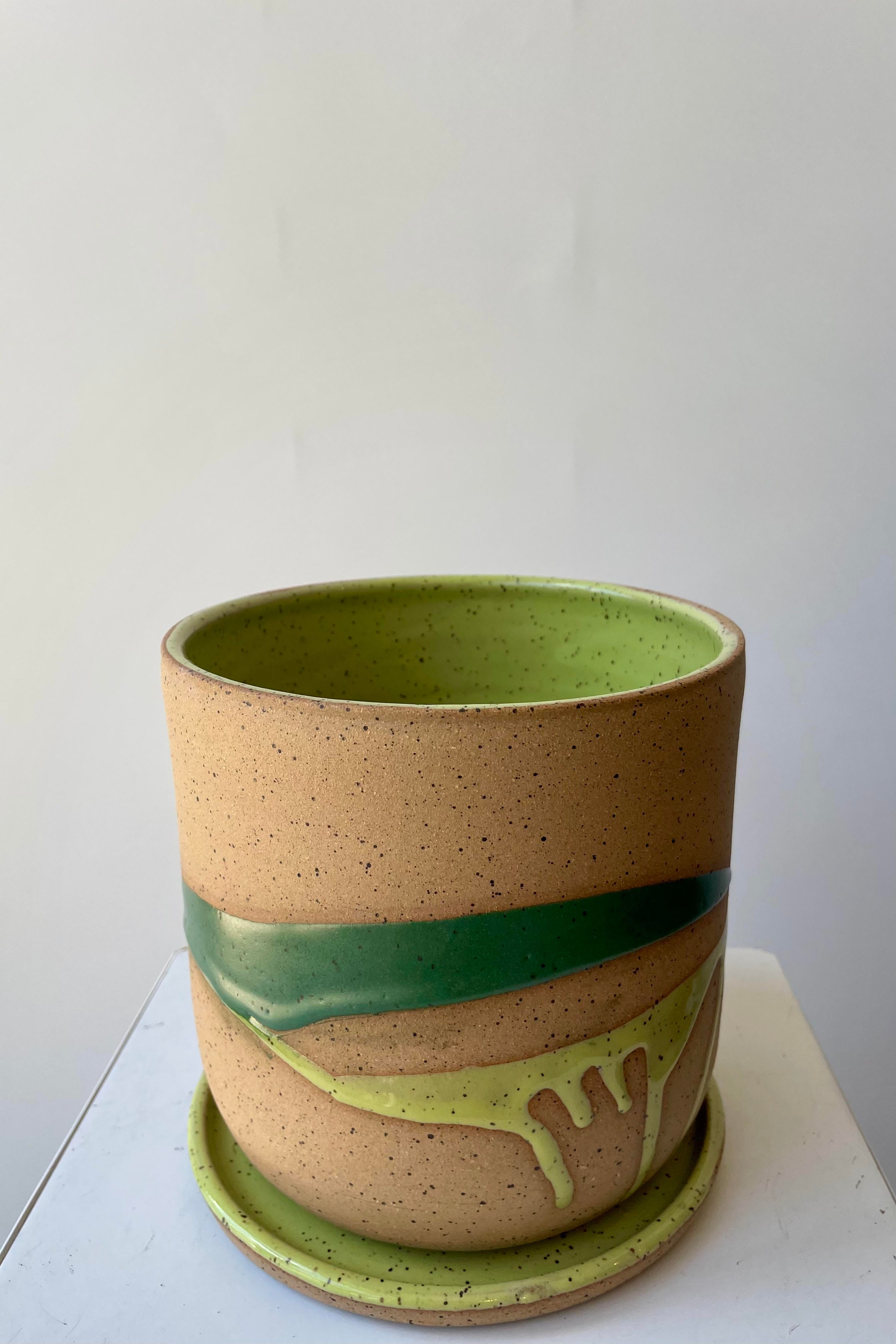 A cylinder plant pot on a saucer. The pot is made of beige clay with a darker speckle pattern. There is a semi-gloss glaze of two shades of green dripped across the planter. The pot is photographed in a white room. The angle of the photo shows the interior green glaze.