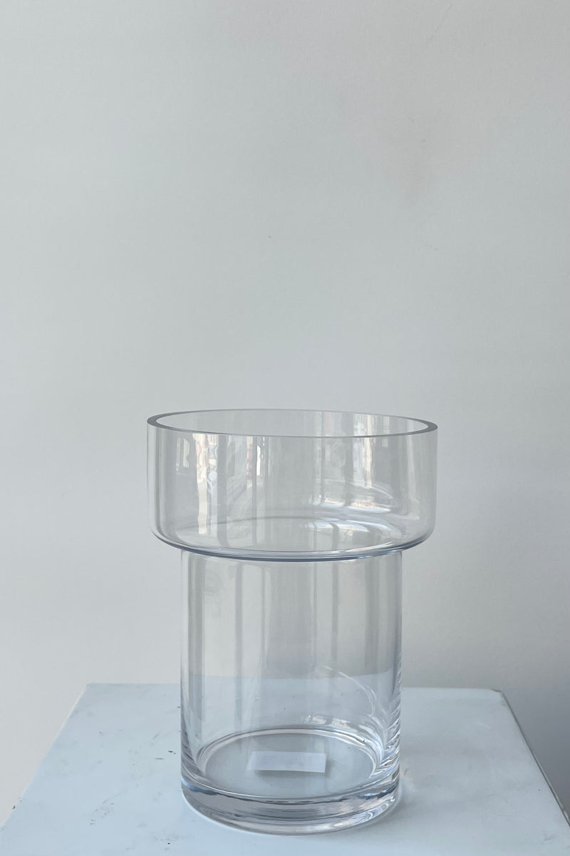 Photo of a clear glass Keeper vase. The vase is cylindrical with a wider top portion and a narrower two-thirds of its height. The vase is photographed in a white room against a white wall.
