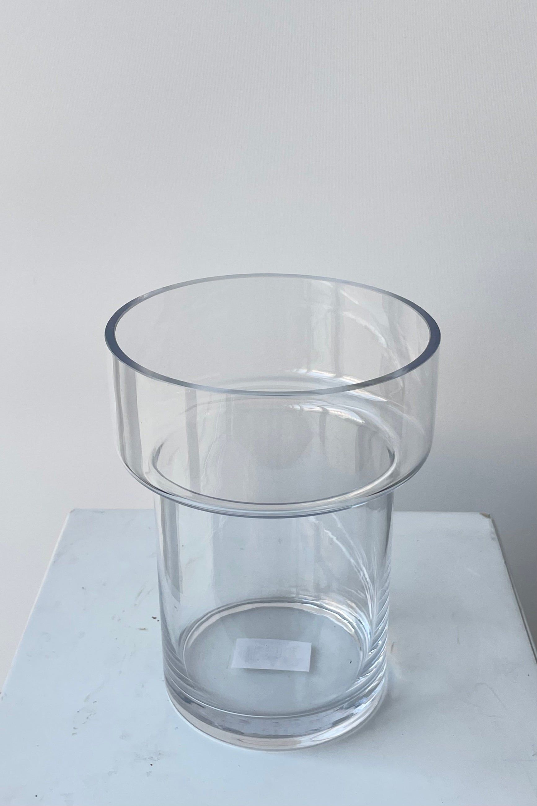 Photo of a clear glass Keeper vase. The vase is cylindrical with a wider top portion and a narrower two-thirds of its height. The vase is photographed in a white room against a white wall. ©Sprout Home