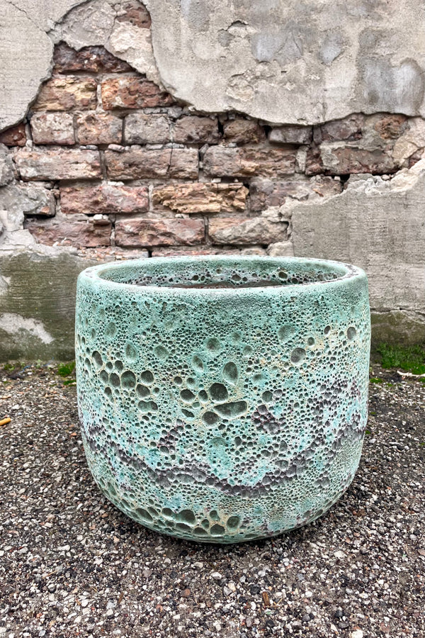 Photo of a Large Coarse Volcano Glaze pot with a heavily textured glaze. Shown outdoors on a paved surface against a brick wall. Shown from the side.