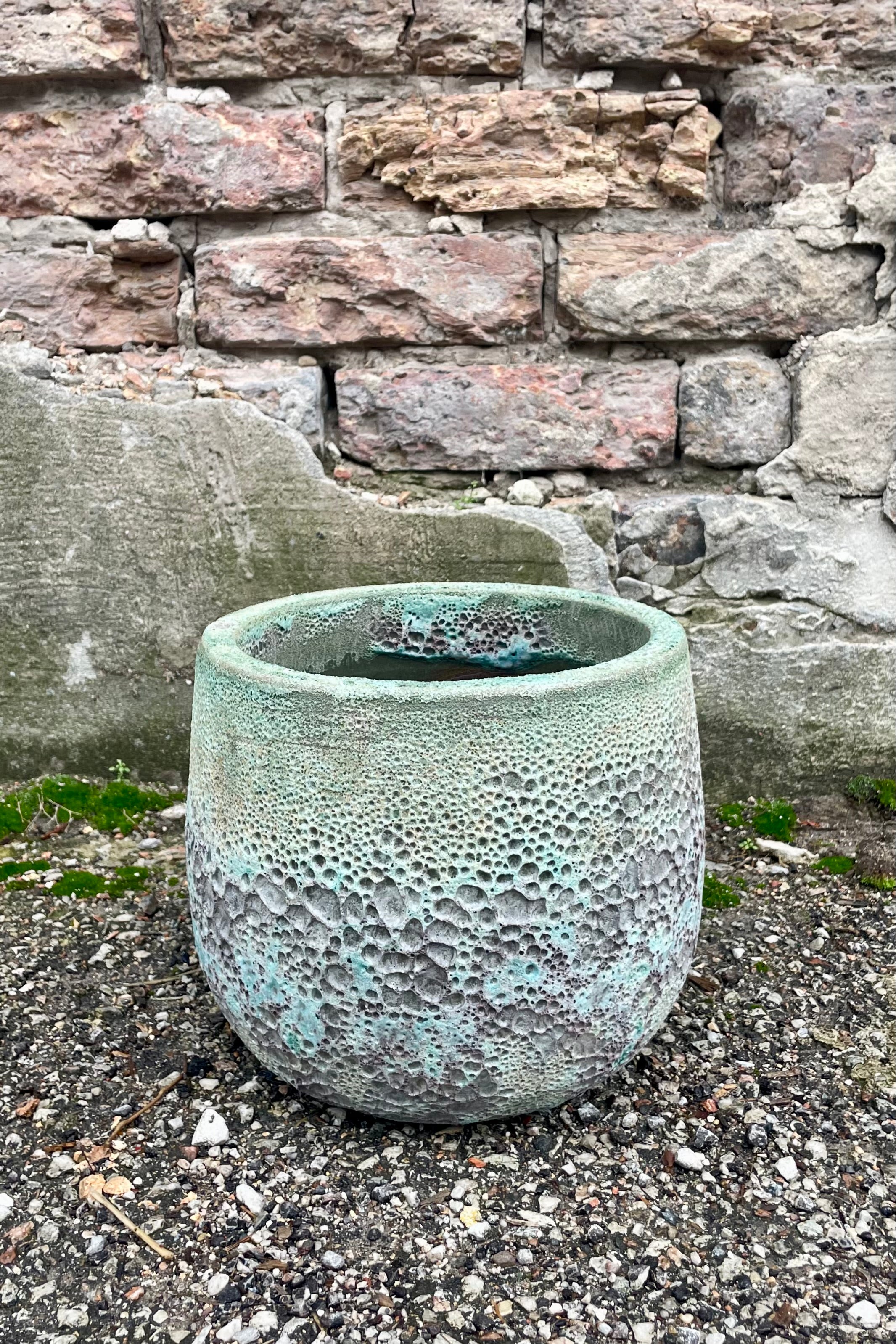 Photo of a Small Coarse Volcano Glaze pot with a heavily textured glaze. Shown outdoors on a paved surface against a brick wall. Shown from the side.