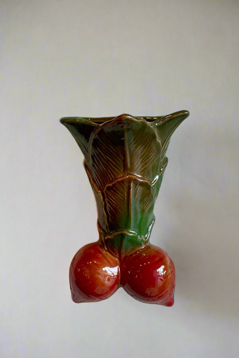 A flower vase hanging on a white wall. The vase hangs on a white wall. The vase is shaped like a pair of red radishes gathered at the leaves that form the body of the vase which is green.