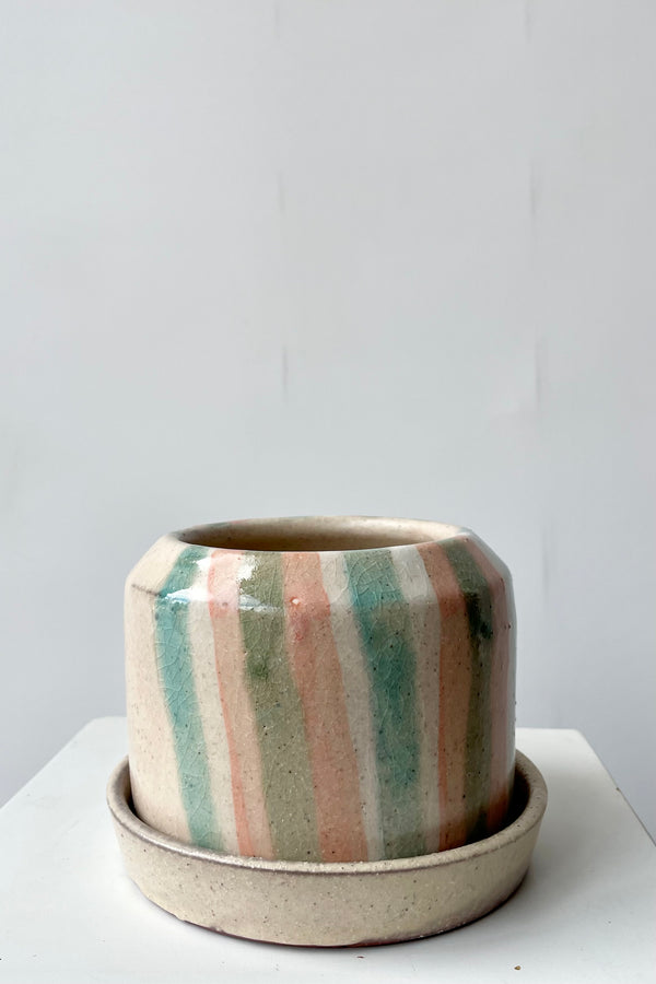 Photo of a glazed terra cotta flower pot and saucer. The planter is set within a saucer and both sit on a white surface in a white room. The semi-gloss glaze features irregular semi-opaque stripes in the glaze. The strapes are shades of coral, light blue and green.
