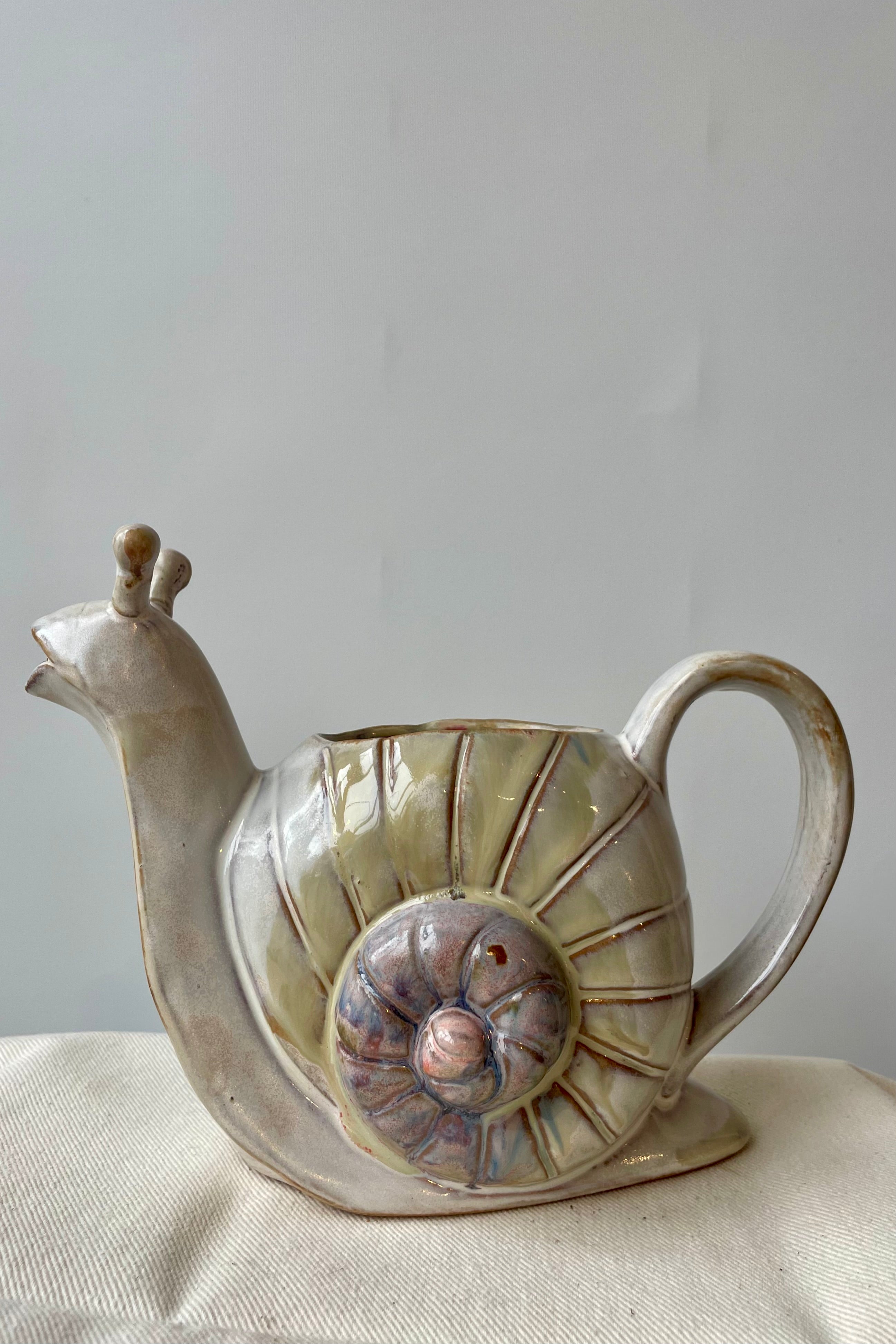 Photo of a stoneware watering can on a white surface against a white wall. The watering can is shaped like a snail and shown from the side showing a spiraled shell with varying color tones along the spiral.