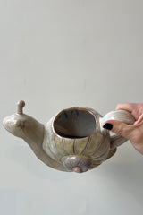 Photo of a stoneware watering can held by a hand in front of a white wall. The watering can is shaped like a snail and shown tipped to the side to show the opening on top.