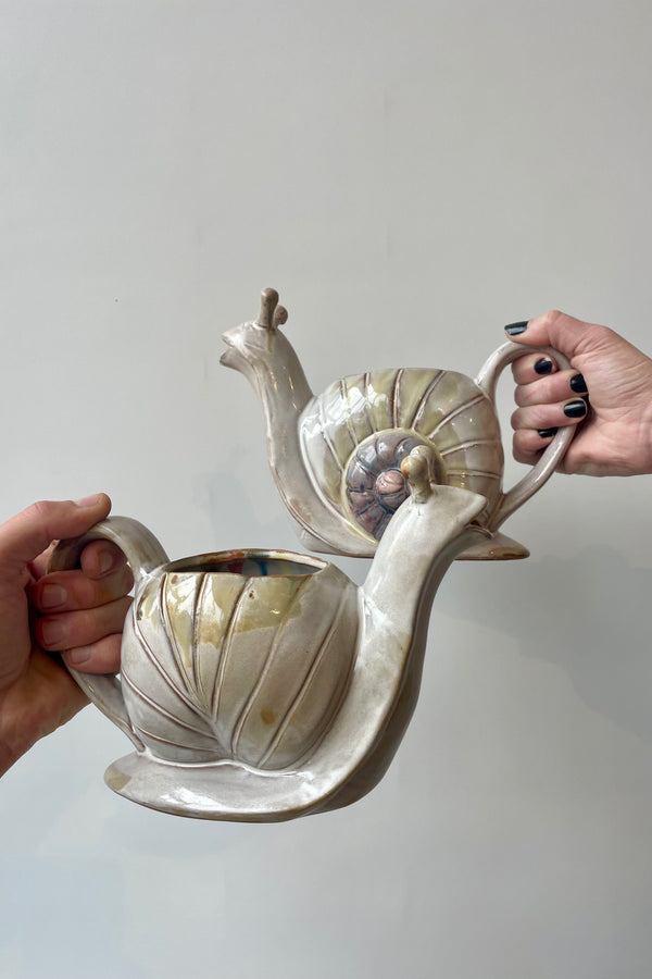 Photo of hands holding stoneware watering cans in front of a white wall. The watering can is shaped like a snail and shown from the side showing a spiraled shell with varying color tones along the spiral on one watering can facing left, and the back of the watering can / shell facing to the right.