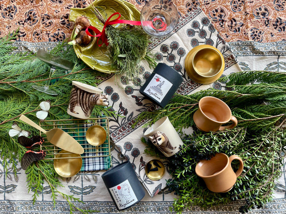 An overhead photo of gift items of brass and ceramic and candles displayed on a bed of evergreen foliage and mixed textiles.