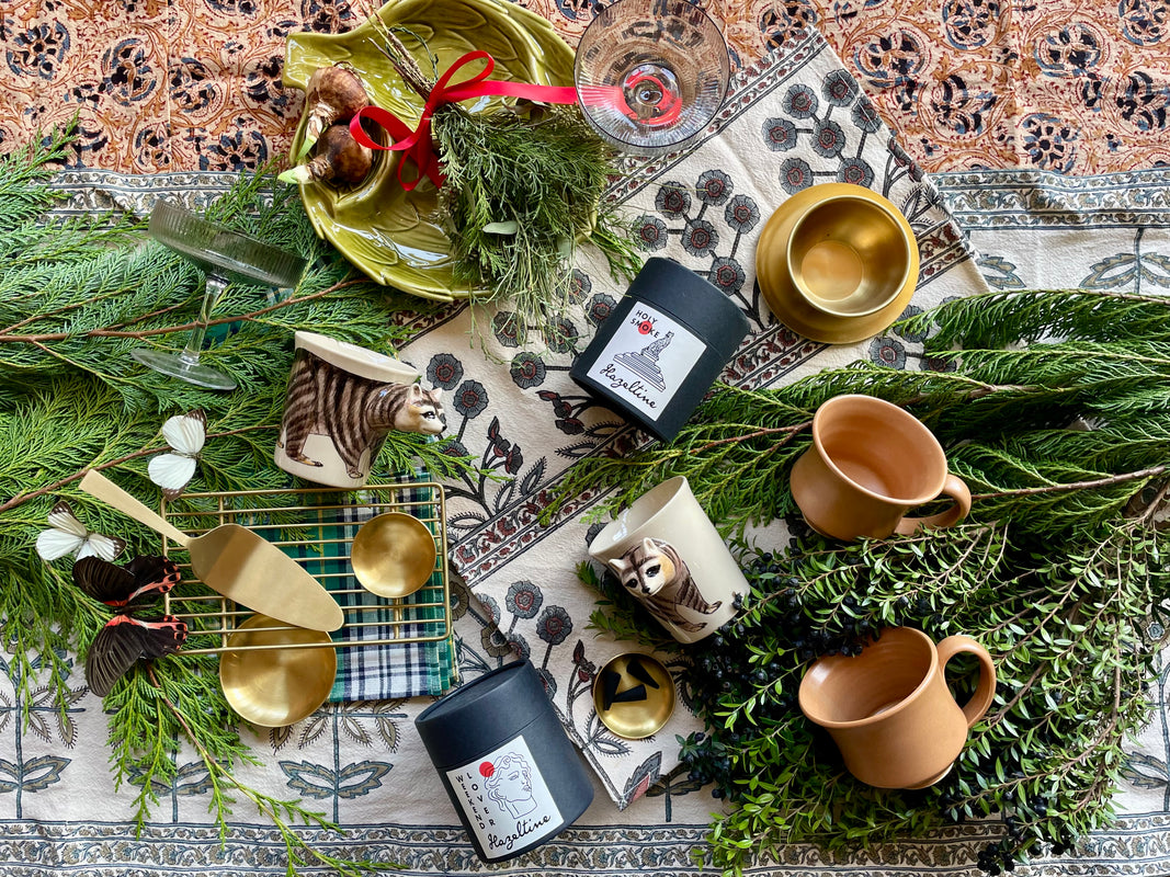 An overhead photo of gift items of brass and ceramic and candles displayed on a bed of evergreen foliage and mixed textiles.