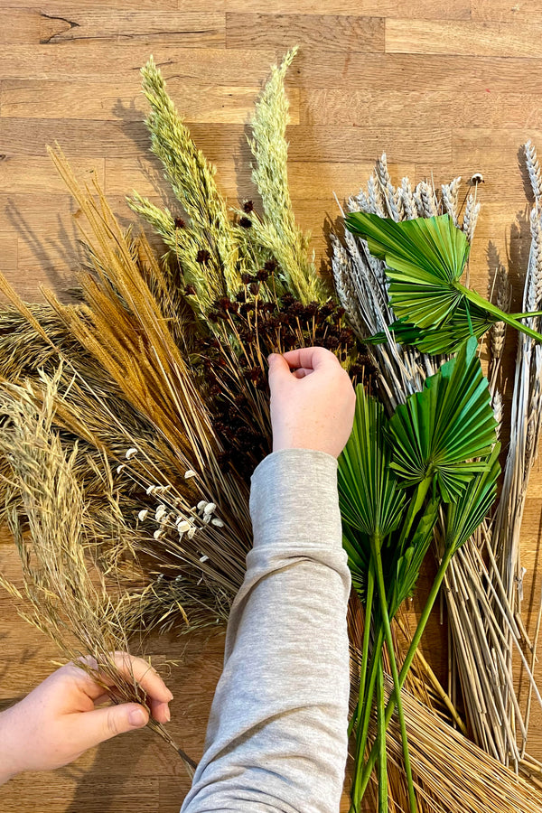 Photo taken from above of hands working with varying textures of green, beige, gold and gray preserved flowers and foliage. They are all placed on a wooden surface.