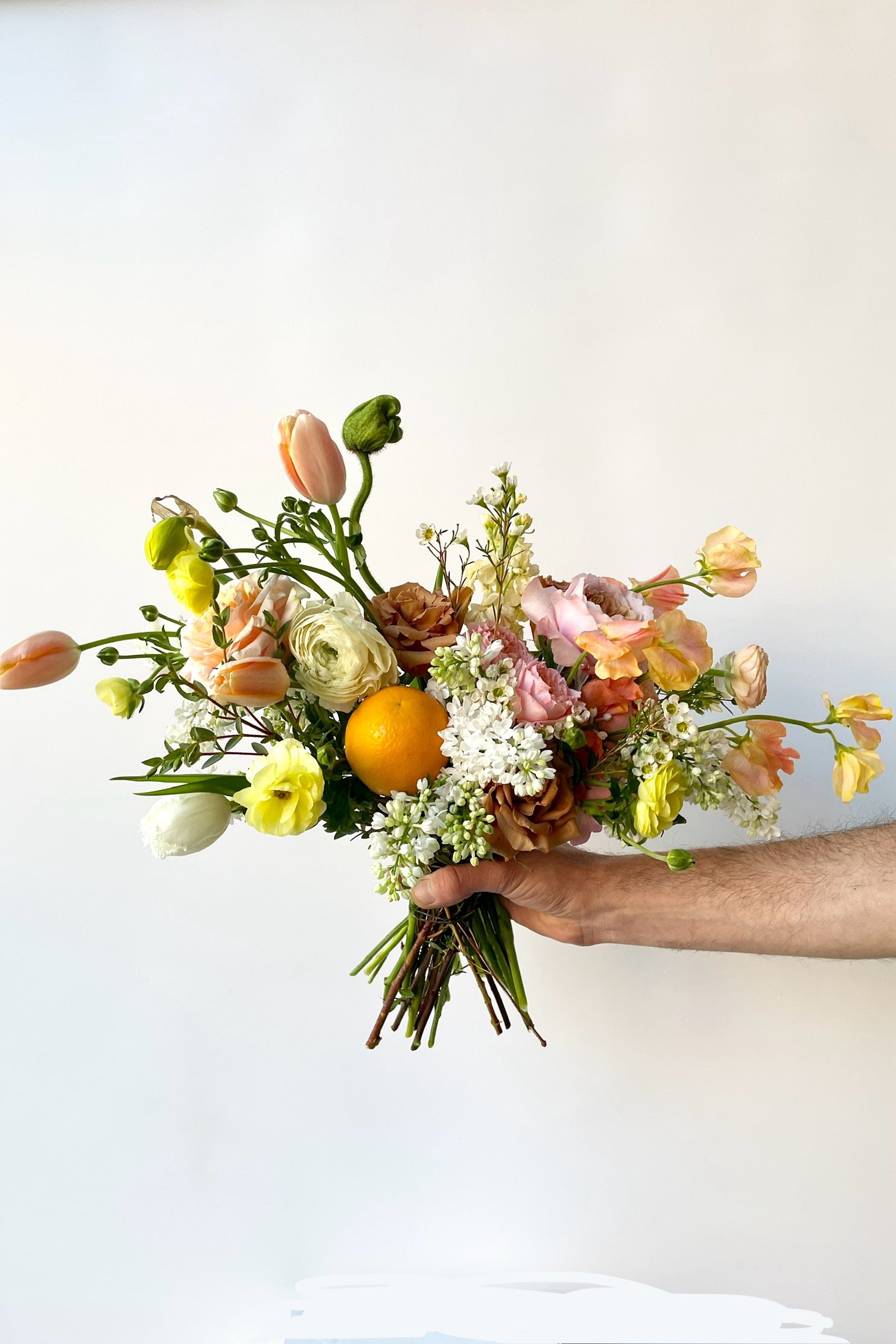 Floral arrangement by SPROUT HOME featuring shades of orange and neutrals, with a real orange being held in hand. Photo by Sprout Home.