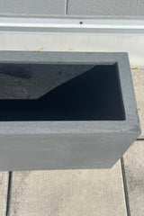 A closer photo from above showing the wide of the the walls of a rectangular planter. The planter is gray and shown against lighter gray brick as a background. The planter is called a Jort planter.