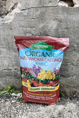 Photo of a plastic bag of Earthworm castings against a brick and cement wall. The bag is maroon with a central band of illustration showing flowers and vegetables as well as the product's name. Below is a golden band with font reading "for organic gardening." The contents and volume are printed at the bottom of the bag.