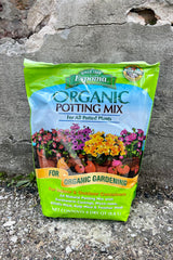 Photo of a plastic bag of organic potting mix against a cement wall. The bag is bright green with a middle band of illustration and text showing the product name and images of flowers and vegetables with the text "for organic gardening." The bottom text shows the contents and volume of the bag.