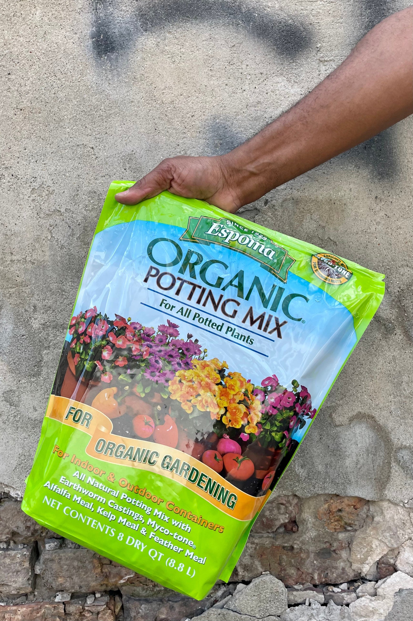 Photo of a hand holding a plastic bag of organic potting mix against a cement wall. The bag is bright green with a middle band of illustration and text showing the product name and images of flowers and vegetables with the text "for organic gardening." The bottom text shows the contents and volume of the bag. ©Sprout Home