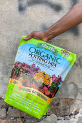 Photo of a hand holding a plastic bag of organic potting mix against a cement wall. The bag is bright green with a middle band of illustration and text showing the product name and images of flowers and vegetables with the text "for organic gardening." The bottom text shows the contents and volume of the bag.