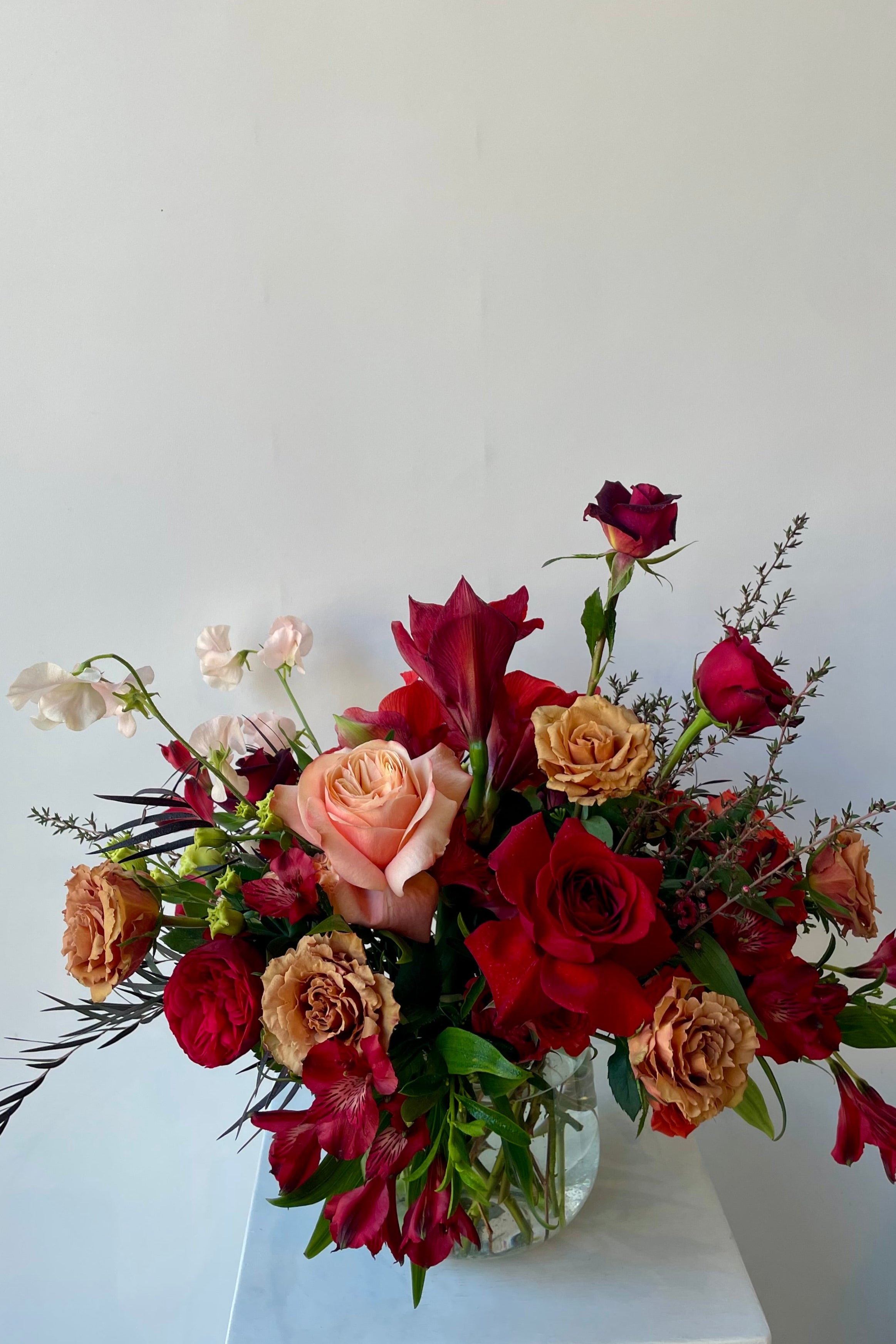 Earth floral arrangement at Sprout Home with Red and Toffee Roses and pink sweet pea styled in clear glass shown against a white wall. #size_$200