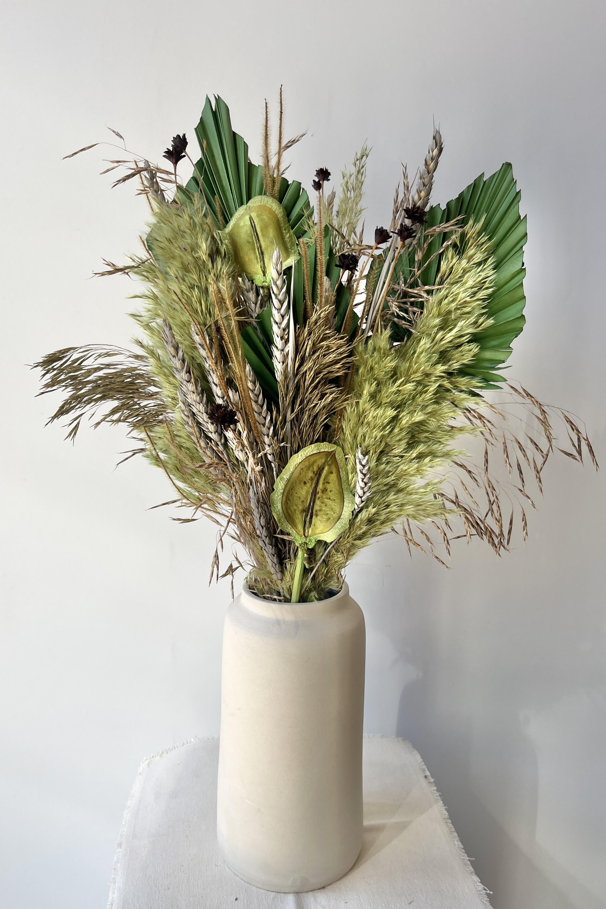 A white vase holding a preserved floral arrangement with a variety of flower types in shades of green, brown, grey and gold against a white wall on a white table. #size_$125