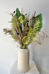 A white vase holding a preserved floral arrangement with a variety of flower types in shades of green, brown, grey and gold against a white wall on a white table.