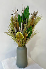 An arrangement of preserved floral and foliage with a variety of flower types in shades of green, brown, grey and gold against a grey wall. The arrangement is shown in a dark green vase.