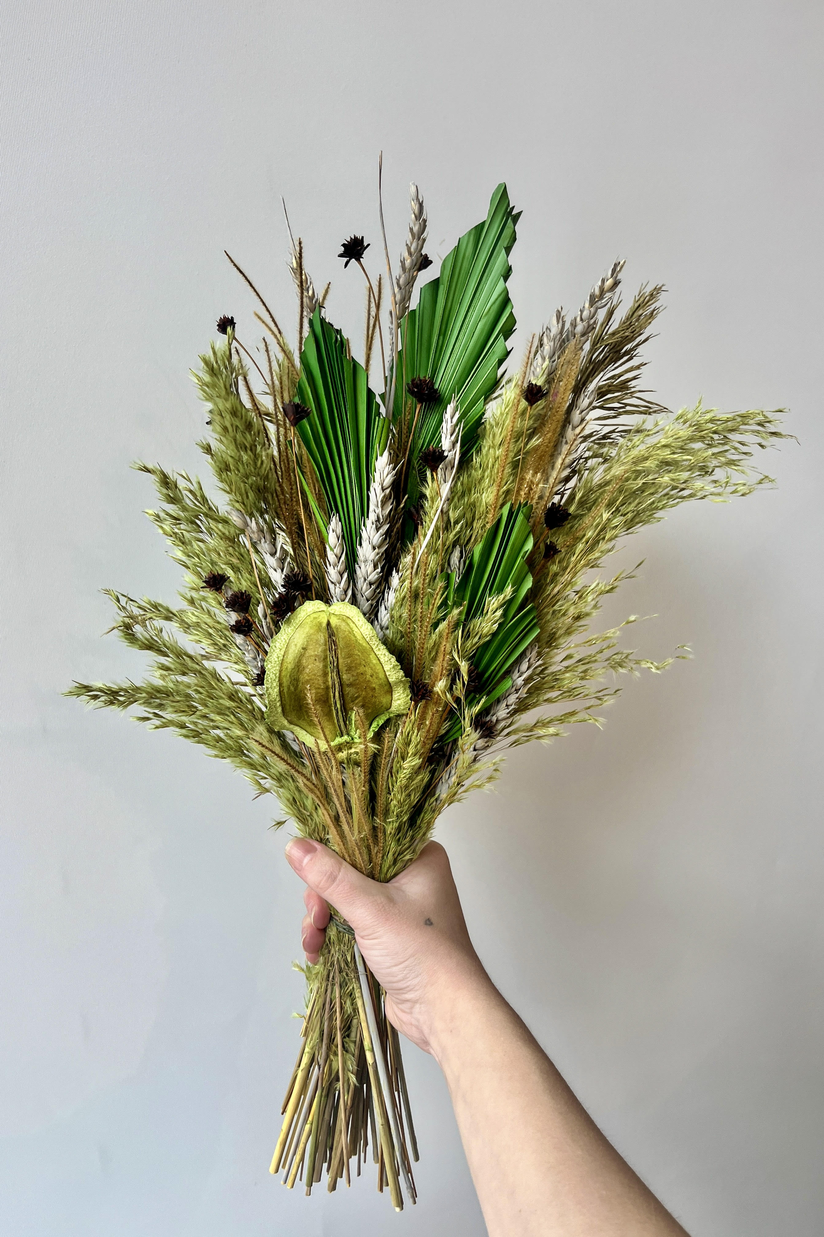 Hand holding preserved floral arrangement with a variety of flower types in shades of green, brown, grey and gold against a white wall. #size_$85