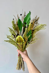 Hand holding preserved floral arrangement with a variety of flower types in shades of green, brown, grey and gold against a white wall. 