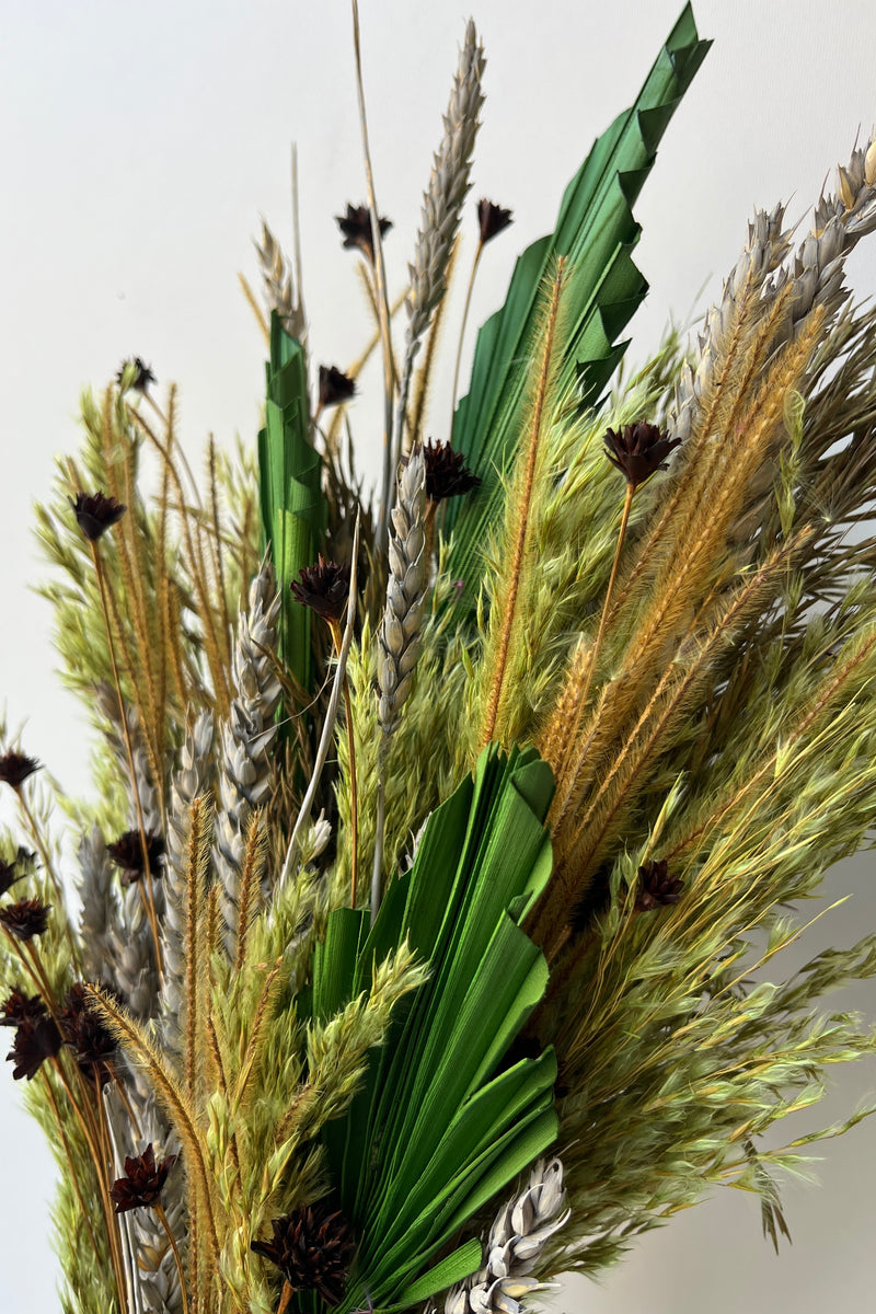 A close up photo of preserved flowers and foliage in varying shades of green, grown, gray and copper. Flowers are shown against a white wall.