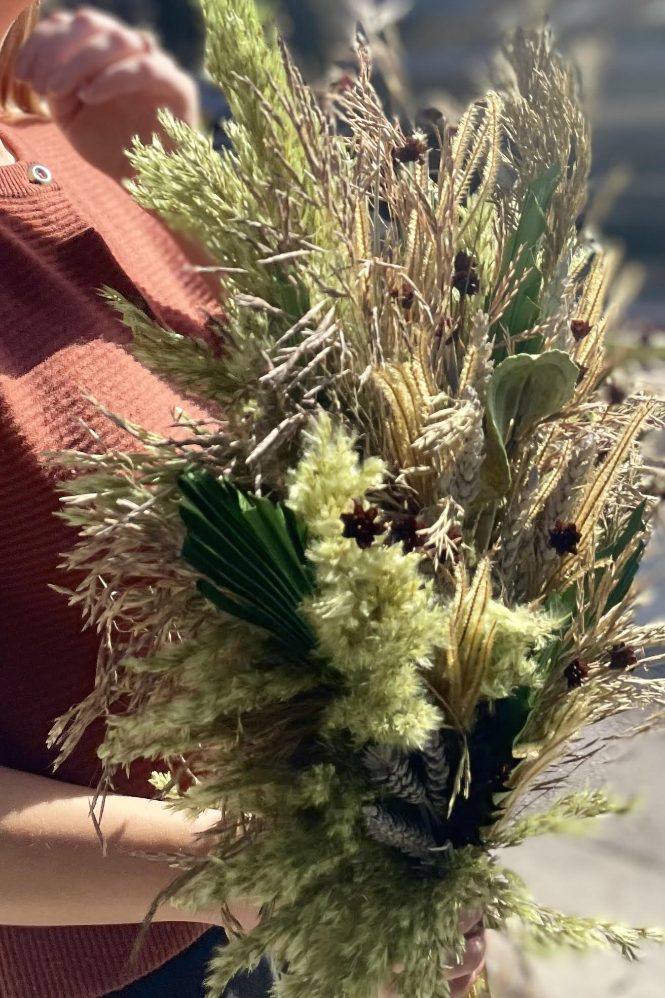 A close photo of a person holding an arrangement of dried flowers and foliage. The flowers are shades of green and brown and beige. Sprout Home #size_$200