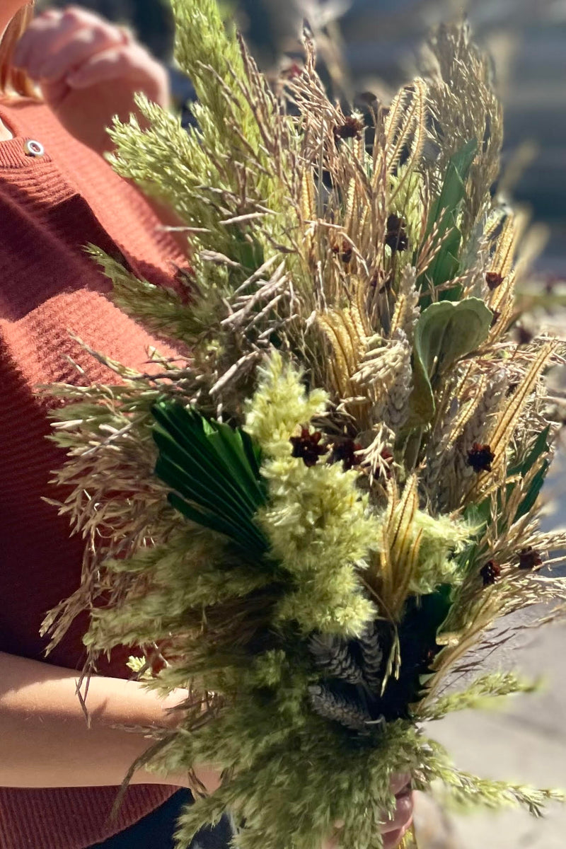 A close photo of a person holding an arrangement of dried flowers and foliage. The flowers are shades of green and brown and beige. 