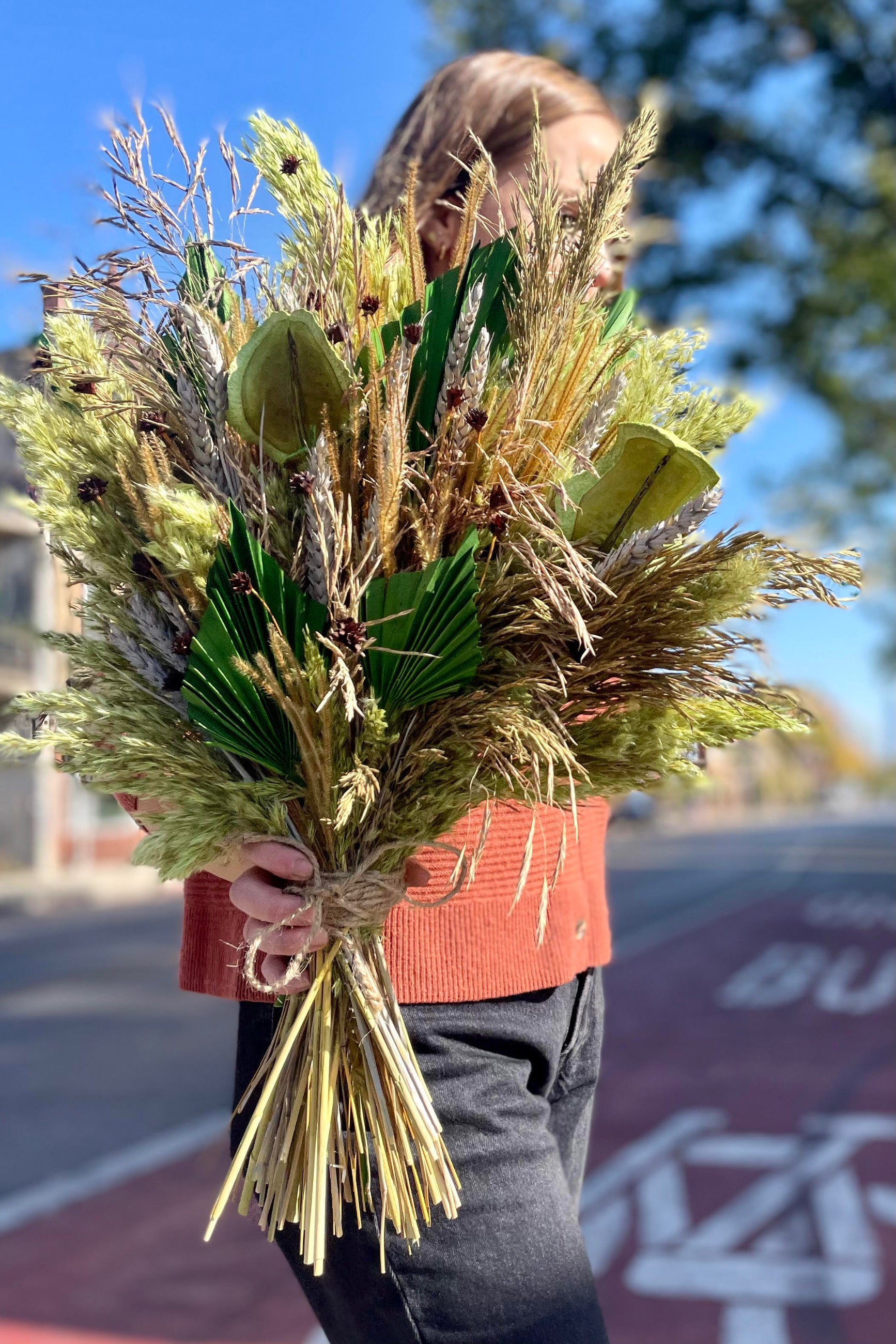 A person holding an arrangement of dried flowers and foliage. The flowers and foliage are shades of green and brown and copper. Their head is turned away from the camper. Blurred buildings are in the background. ©Sprout Home #size_$200