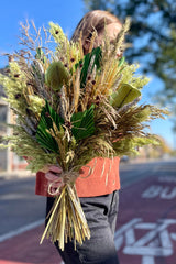 A person holding an arrangement of dried flowers and foliage. The flowers and foliage are shades of green and brown and copper. Their head is turned away from the camper. Blurred buildings are in the background.