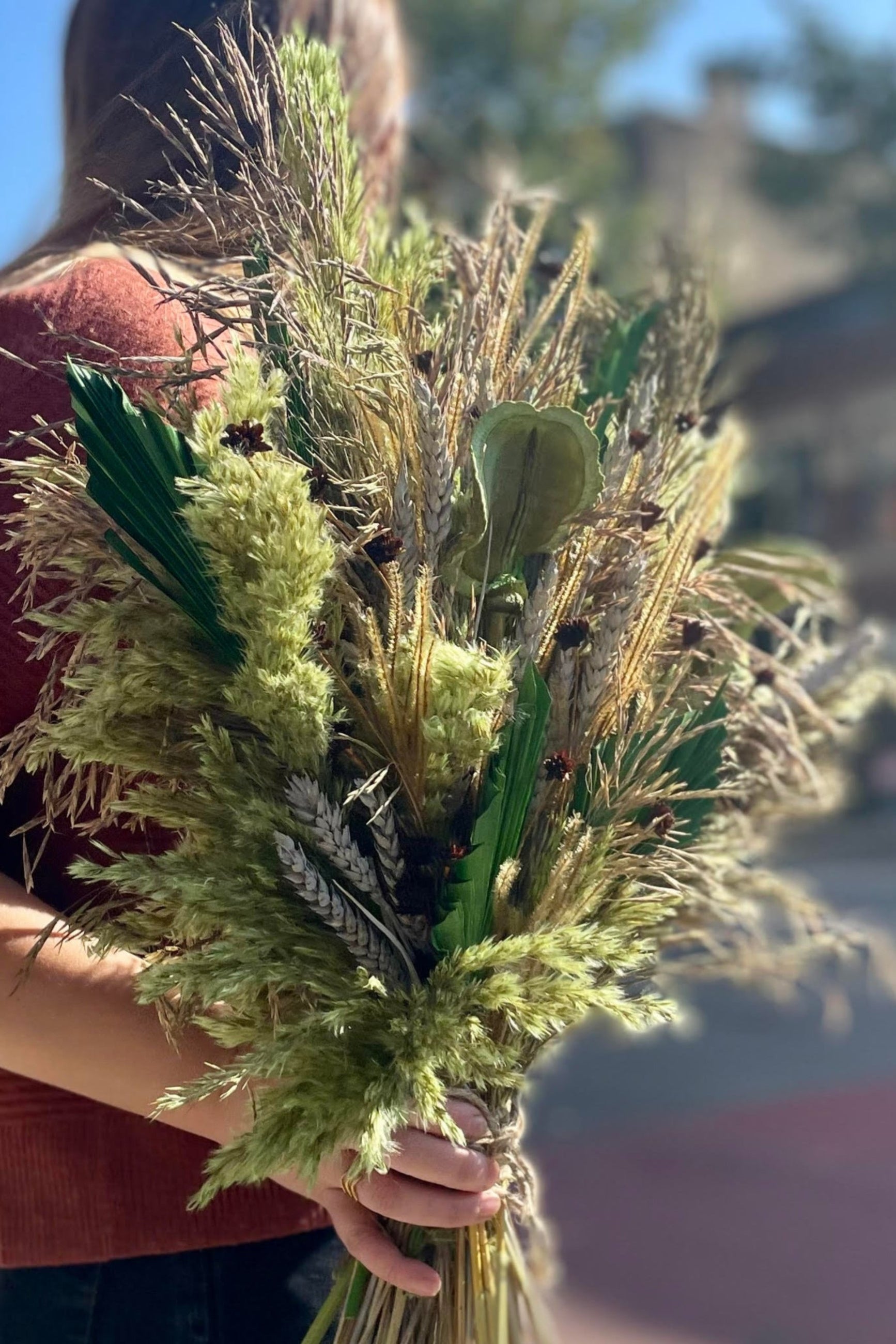 A person holding an arrangement of dried flowers and foliage. The flowers and foliage are shades of green and brown and copper. Their head is turned away from the camper. Blurred buildings are in the background. ©Sprout Home #size_PLP