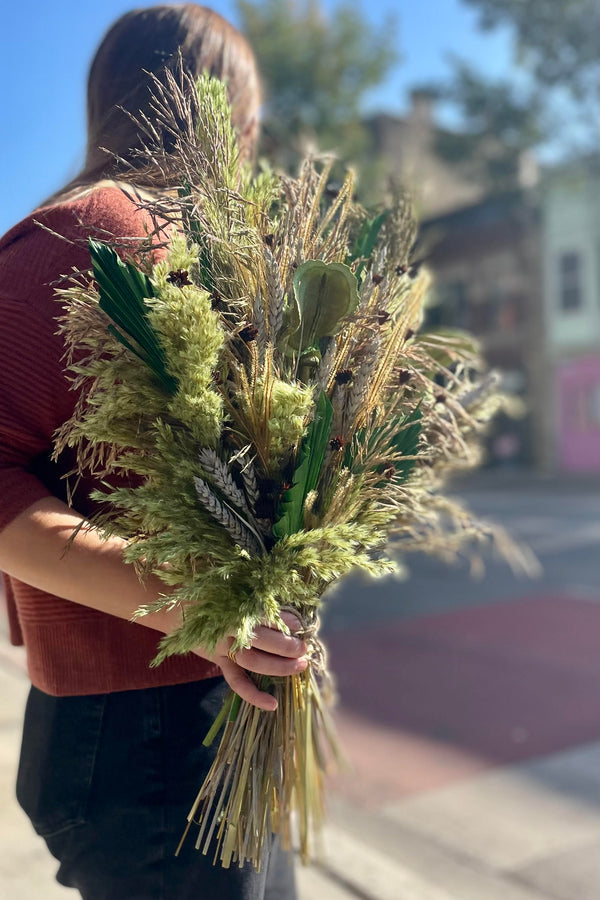 A person holding an arrangement of dried flowers and foliage. The flowers and foliage are shades of green and brown and copper. Their head is turned away from the camper. Blurred buildings are in the background.