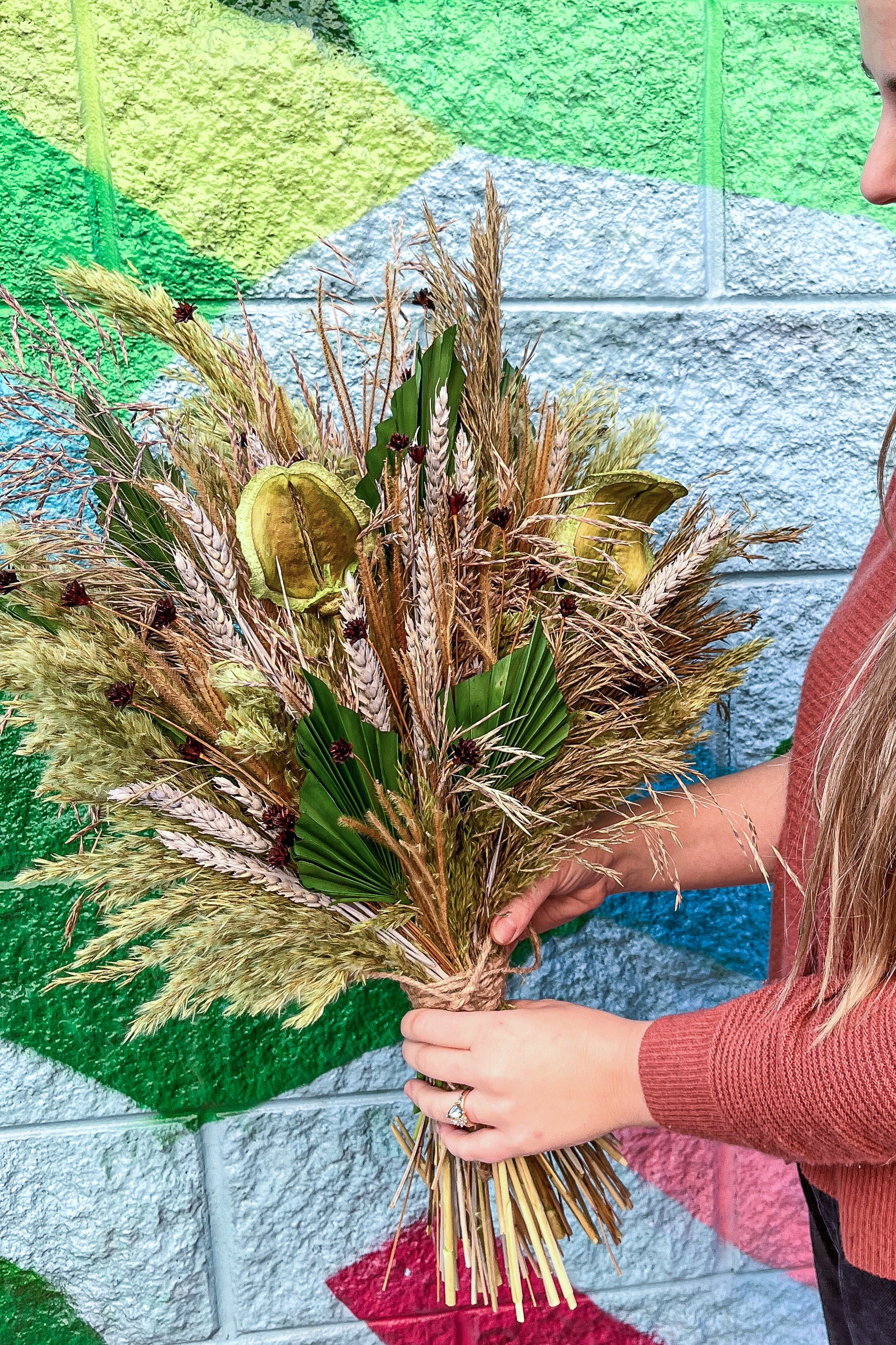 A photo of a person holding an arrangement of dried flowers and foliage. The flowers are shades of green and brown and beige. #size_$200