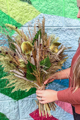 A photo of a person holding an arrangement of dried flowers and foliage. The flowers are shades of green and brown and beige. 