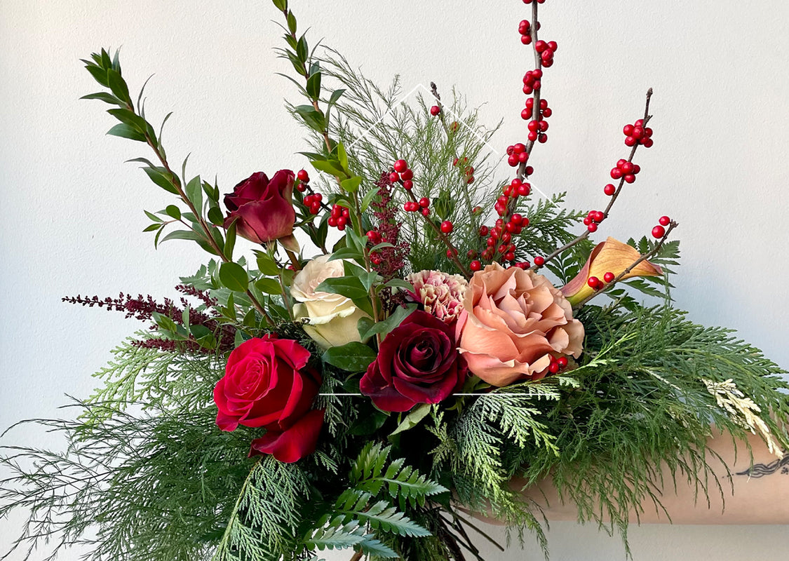 Fresh floral arrangement with red roses, winterberry, cedar, boxwood and peach accent flowers against a white wall.