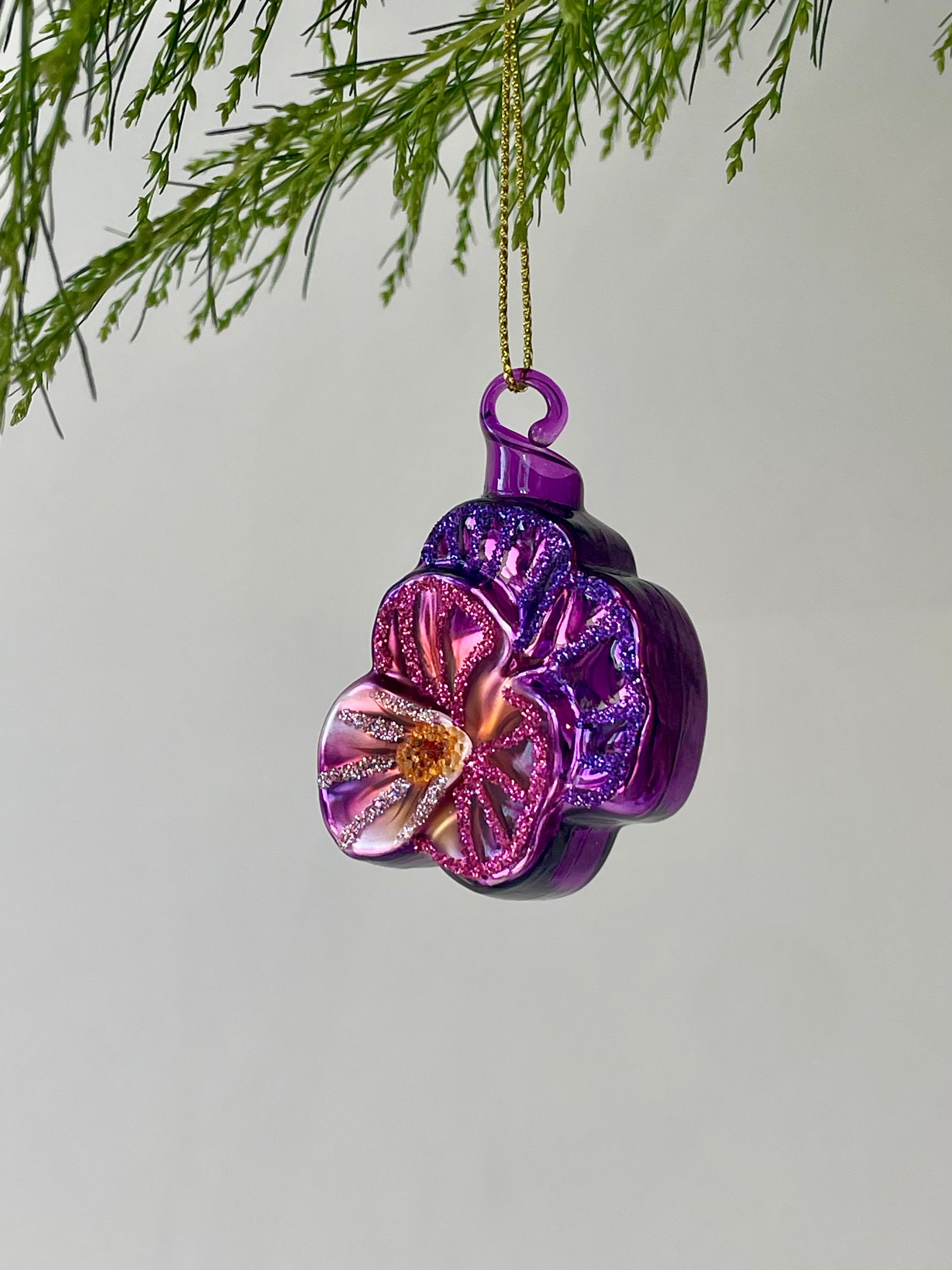 A purple floral ornament shaped like a violet with glitter hangs from a branch in front of a gray wall ©Sprout Home