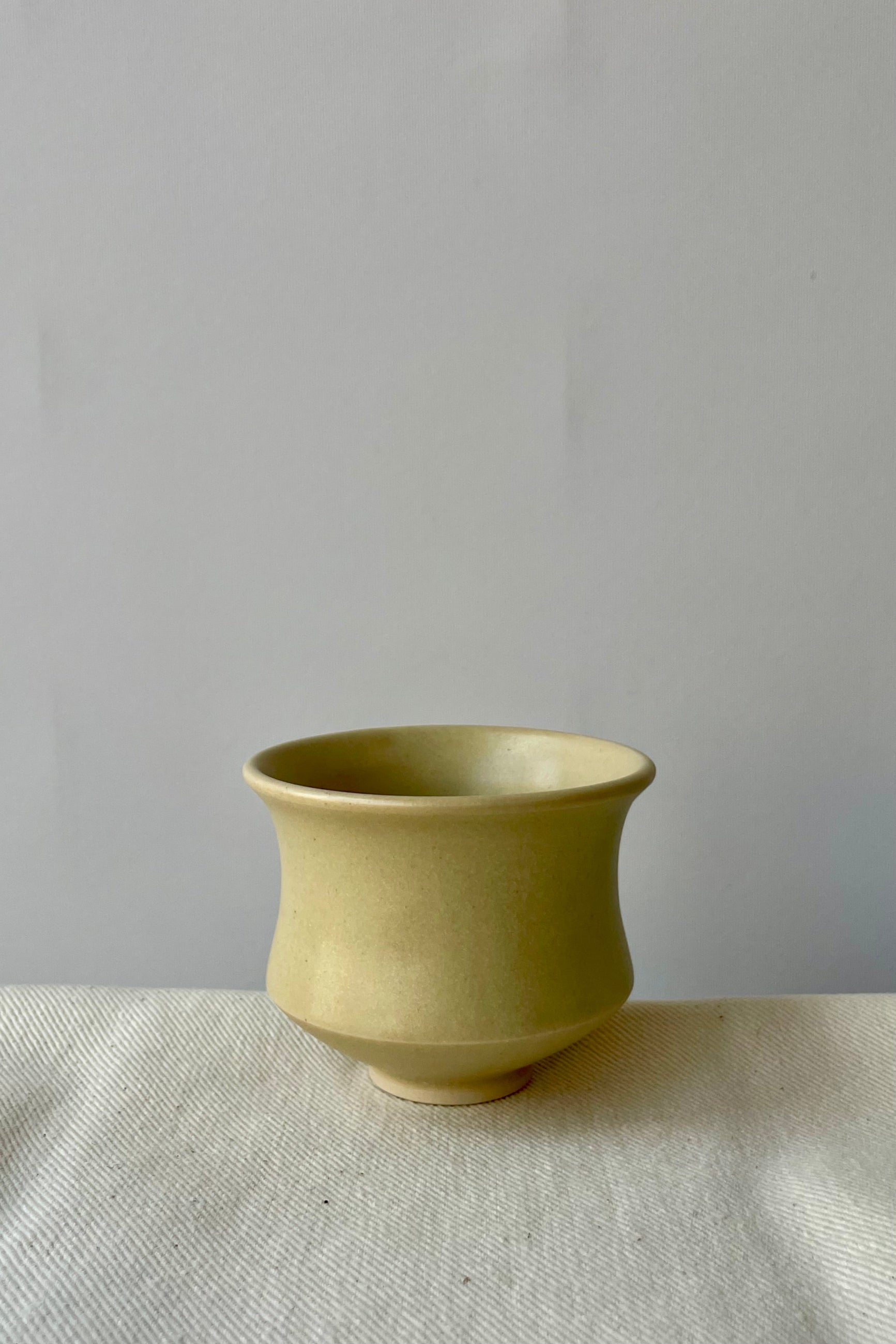 Photo of Little Fire Ceramics hourglass cup with butter glaze shown from the side. The cup sits on a white cloth against a white wall. ©Sprout Home