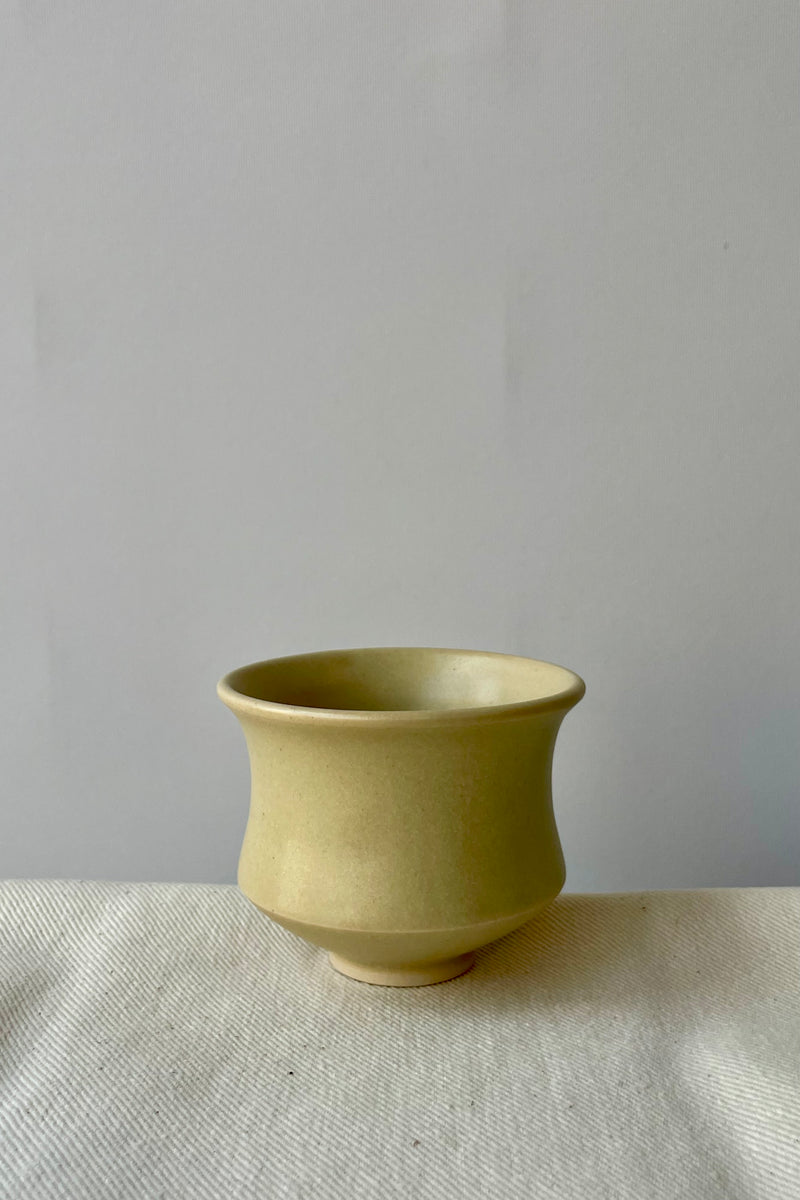 Photo of Little Fire Ceramics hourglass cup with butter glaze shown from the side. The cup sits on a white cloth against a white wall.