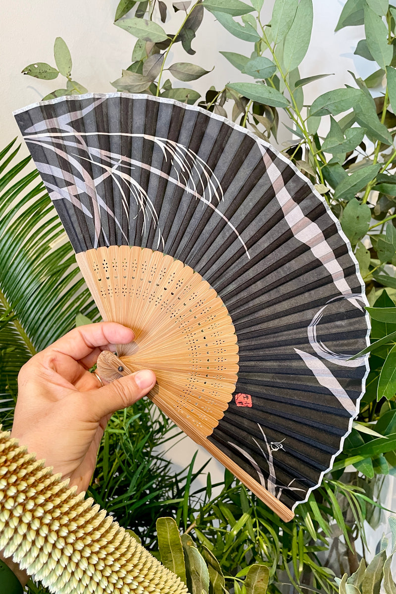 A black and grey fan being held in hand in front of foliage at Sprout Home