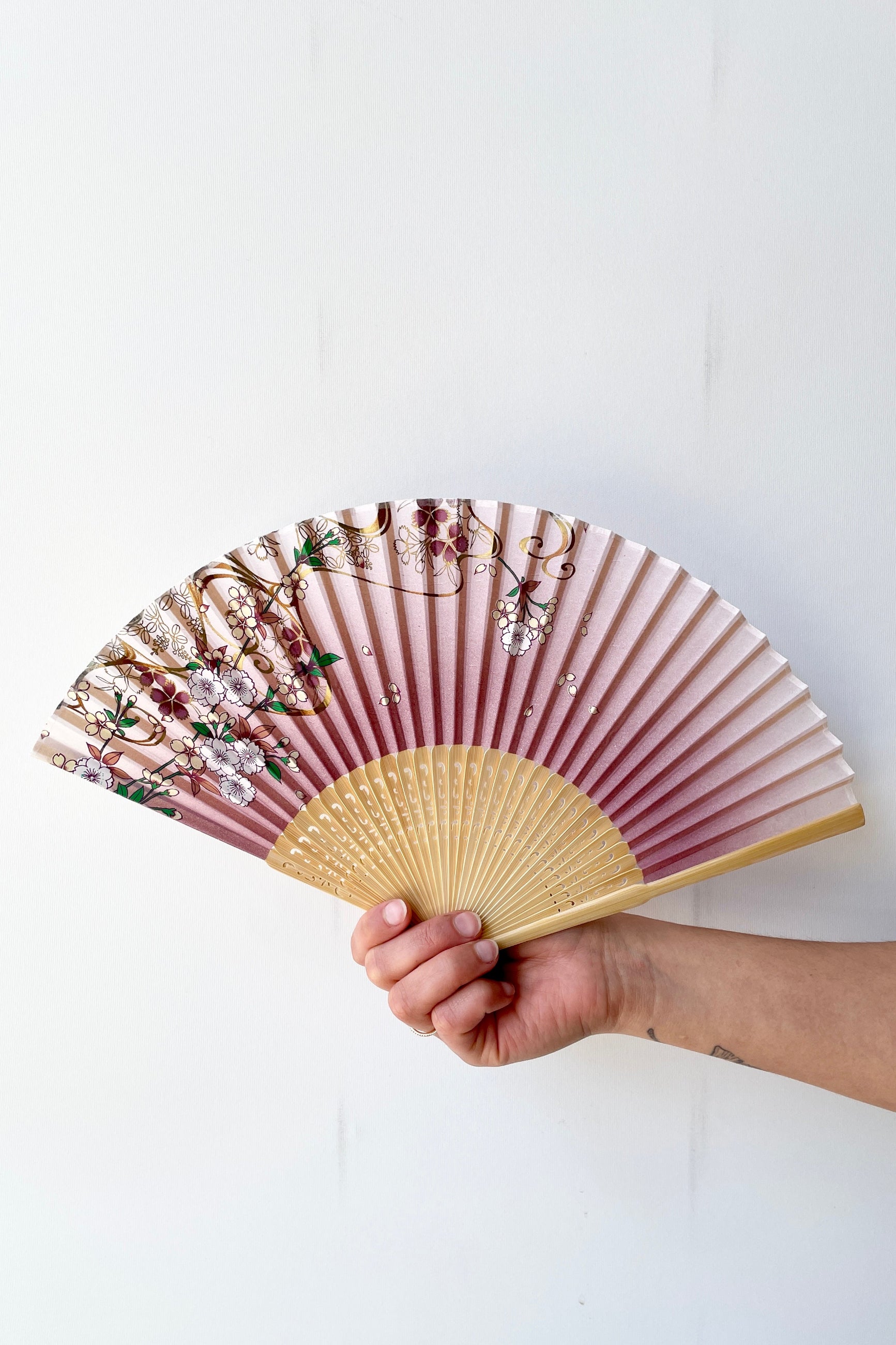 Silk fan cherry blossom pattern with gold being held open in hand against a white wall ©Sprout Home